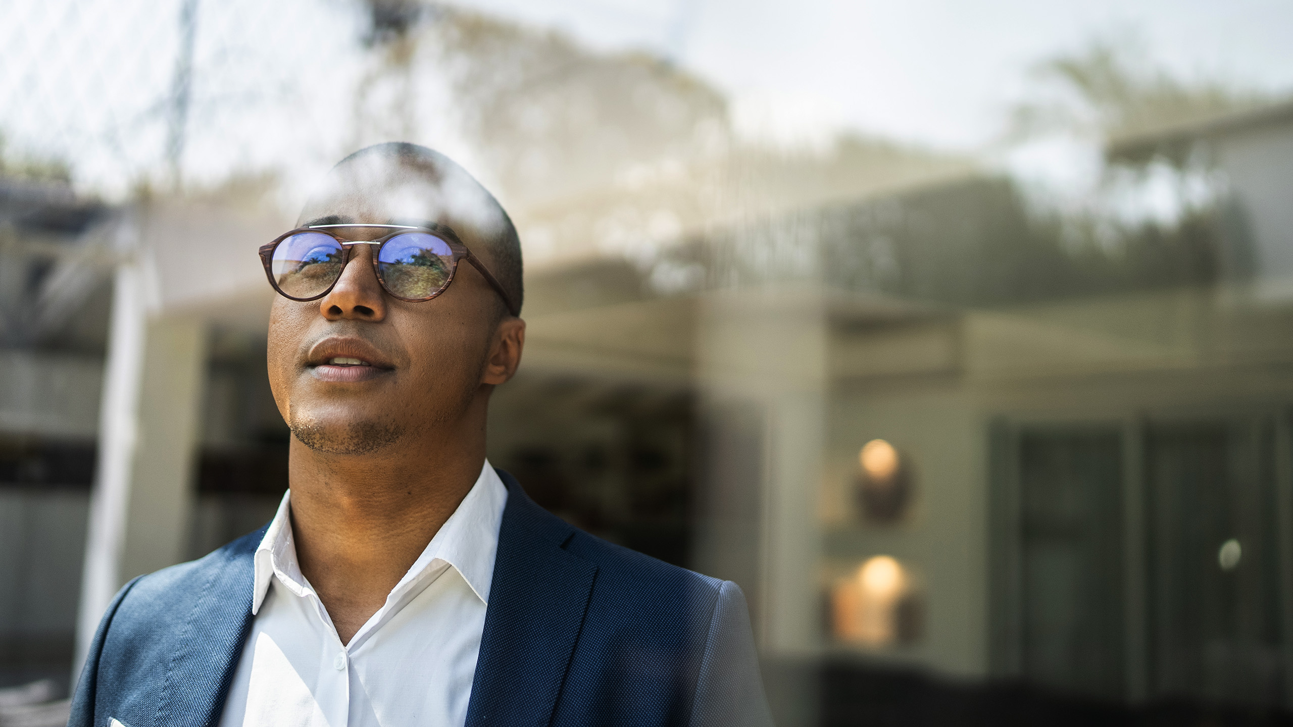 Personne portant une tenue professionnelle debout à l’extérieur près d’une grande fenêtre, avec des reflets de l’environnement visibles sur la surface vitrée.